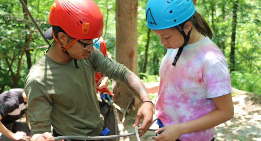 An outward bound instructor helps a student tie a rope to their harness. They are both wearing helmets and stand in a wooded area. 